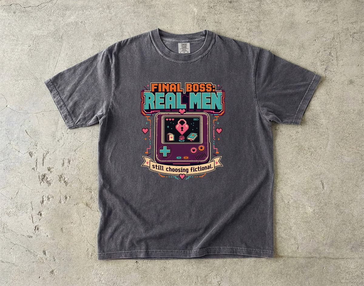 A dark gray graphic t-shirt laid flat on a textured concrete surface. The shirt features a retro video game console graphic with the text 'FINAL BOSS REAL MEN' and 'still choosing fictionals'.
