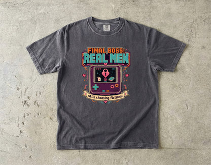 A dark gray graphic t-shirt laid flat on a textured concrete surface. The shirt features a retro video game console graphic with the text 'FINAL BOSS REAL MEN' and 'still choosing fictionals'.