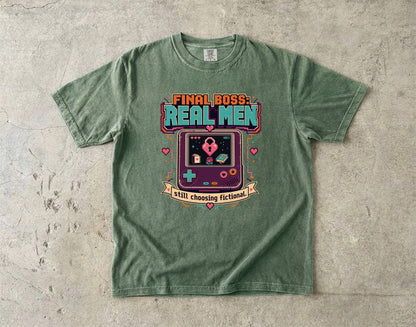 A green graphic t-shirt laid flat on a textured concrete surface. The shirt features a retro video game console graphic with the text 'FINAL BOSS REAL MEN' and 'Still choosing fictional!' in colorful lettering.