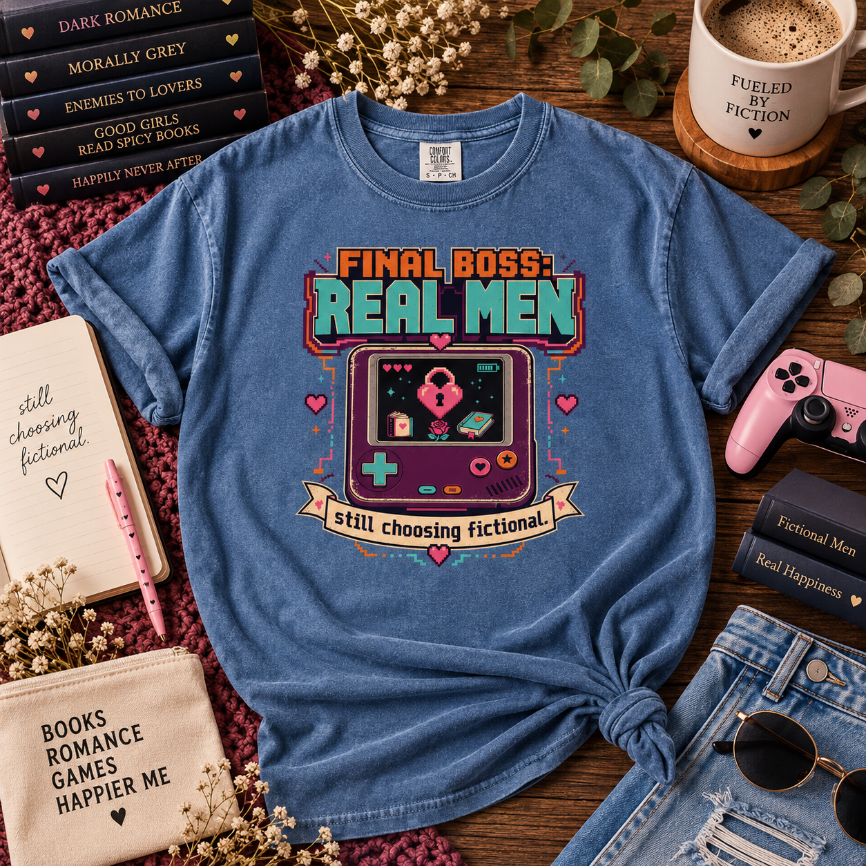 A blue graphic t-shirt featuring a retro video game design with the text 'FINAL BOSS REAL MEN' and 'still choosing fictional.' styled in a cozy flat-lay arrangement with books, a pink game controller, sunglasses, and a mug on a wooden surface.