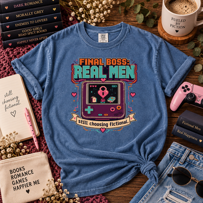 A blue graphic t-shirt featuring a retro video game design with the text 'FINAL BOSS REAL MEN' and 'still choosing fictional.' styled in a cozy flat-lay arrangement with books, a pink game controller, sunglasses, and a mug on a wooden surface.
