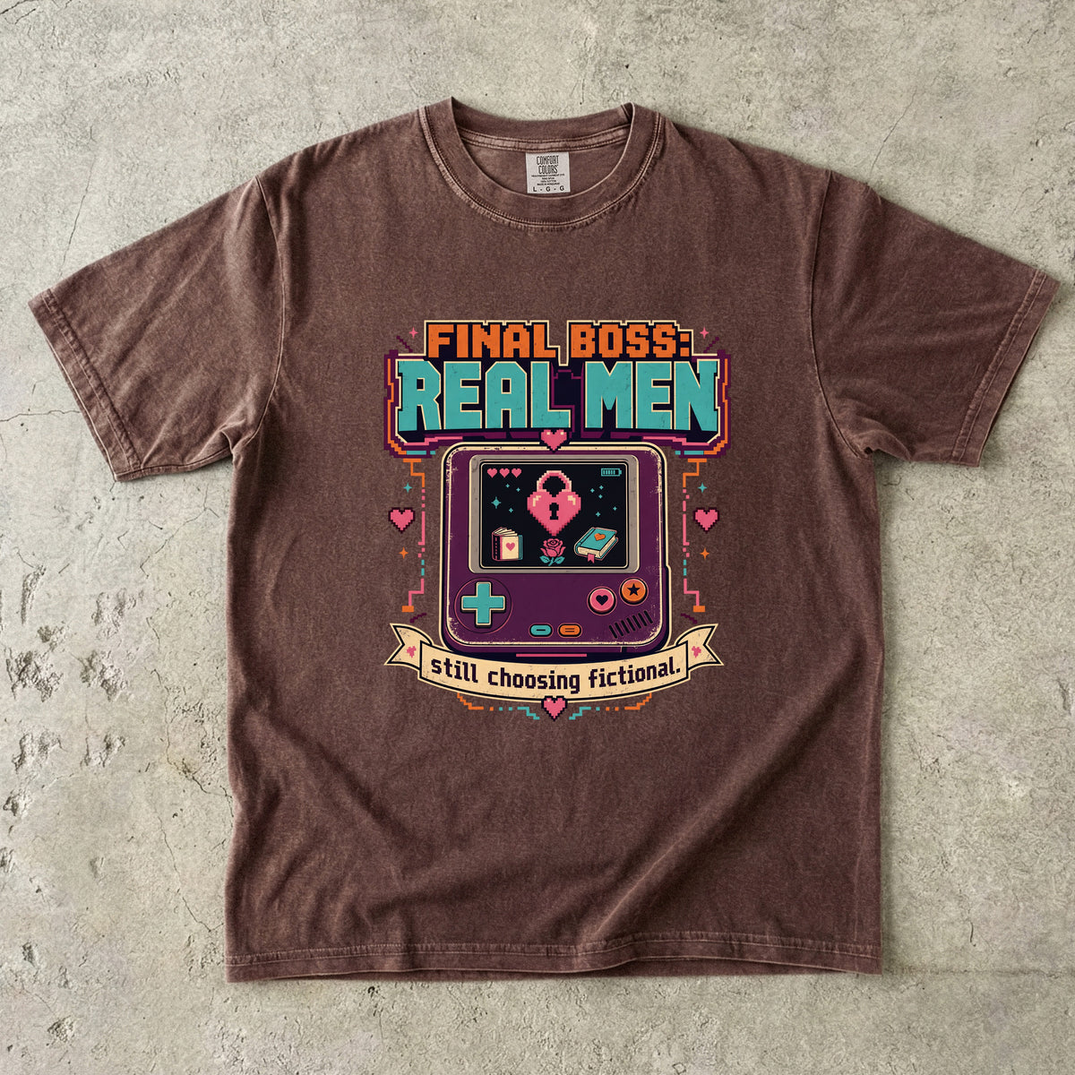 A brown short-sleeve t-shirt with a colorful retro video game graphic on the chest, laid flat on a textured gray concrete surface. The graphic features the text 'FINAL BOSS REAL MEN' and 'still choosing fictional' around a retro handheld game console illustration.