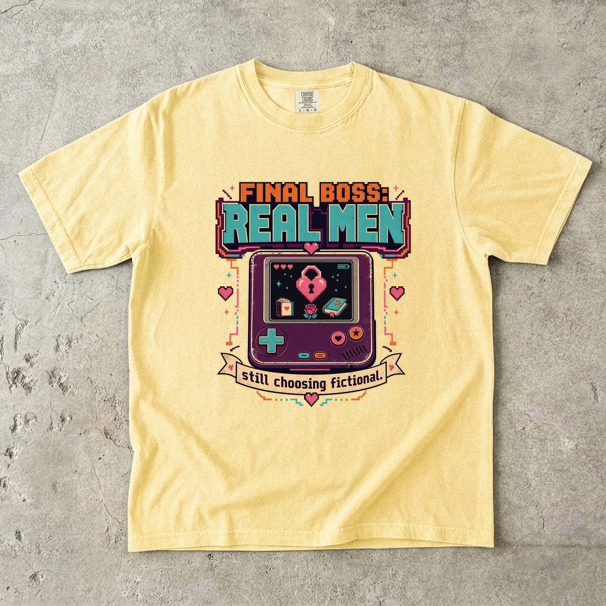 A pale yellow graphic t-shirt laid flat on a textured gray concrete surface. The shirt features a retro video game handheld console graphic with the text 'FINAL BOSS: REAL MEN' and 'I still choosing fictional.