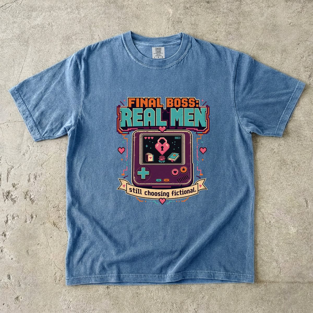 A light blue crewneck t-shirt laid flat on a textured concrete surface, featuring a retro video game handheld console graphic with the text 'FINAL BOSS: REAL MEN' and 'still choosing fictional!'.