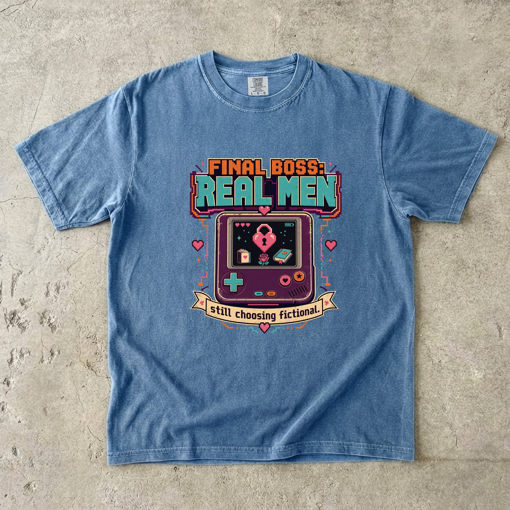 A light blue crewneck t-shirt laid flat on a textured concrete surface, featuring a retro video game handheld console graphic with the text 'FINAL BOSS: REAL MEN' and 'still choosing fictional!'.