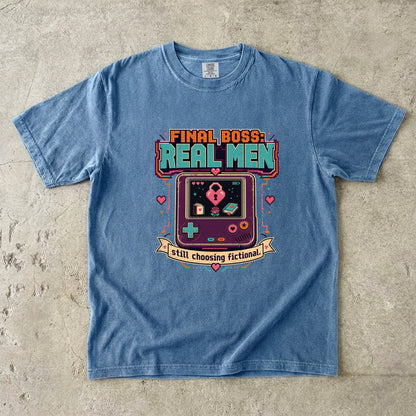 A light blue crewneck t-shirt laid flat on a textured concrete surface, featuring a retro video game handheld console graphic with the text 'FINAL BOSS: REAL MEN' and 'still choosing fictional!'.