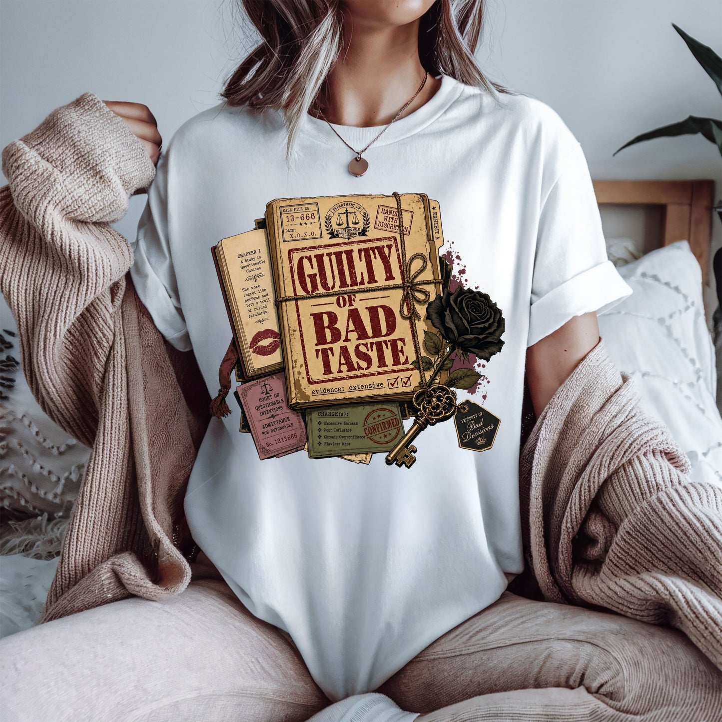 A woman wearing a white crewneck t-shirt with a vintage-style graphic print of a book titled 'Guilty of Bad Taste' tied with a rope and decorated with a black rose and key. She is wearing a beige knit cardigan draped off her shoulders in a cozy bedroom setting.