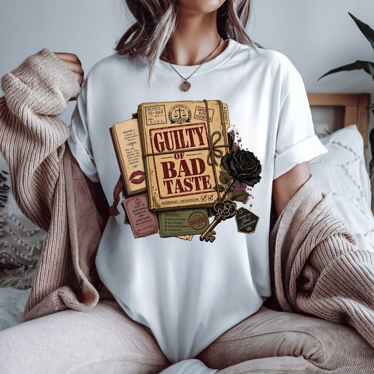 A woman wearing a white crewneck t-shirt with a vintage-style graphic print of a book titled 'Guilty of Bad Taste' tied with a rope and decorated with a black rose and key. She is wearing a beige knit cardigan draped off her shoulders in a cozy bedroom setting.