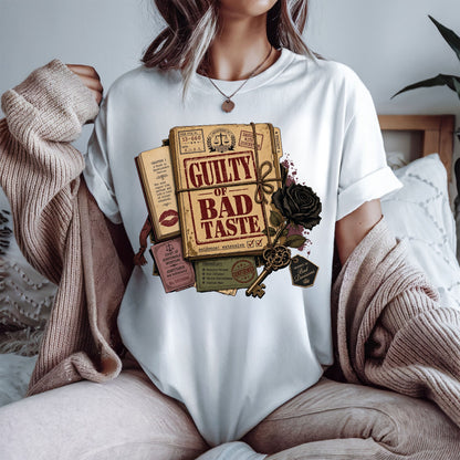 A woman wearing a white crewneck t-shirt with a vintage-style graphic print of a book titled 'Guilty of Bad Taste' tied with a rope and decorated with a black rose and key. She is wearing a beige knit cardigan draped off her shoulders in a cozy bedroom setting.