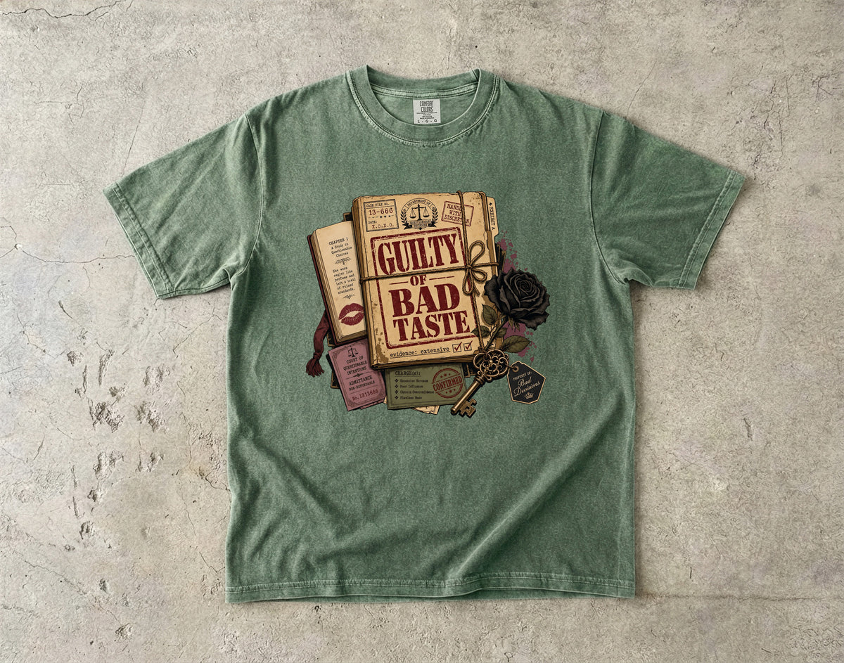 A sage green crewneck t-shirt laid flat on a textured concrete surface, featuring a graphic print of vintage books, a key, and a black rose with the text 'GUILTY BAD TASTE'.