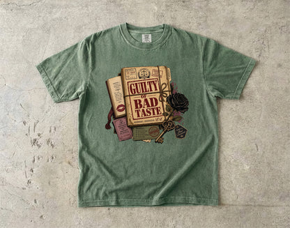 A sage green crewneck t-shirt laid flat on a textured concrete surface, featuring a graphic print of vintage books, a key, and a black rose with the text 'GUILTY BAD TASTE'.