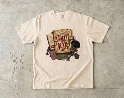 A cream-colored short-sleeve t-shirt laid flat on a textured gray concrete surface. The front of the t-shirt features a graphic print of vintage books, a black rose, and a key with the text 'GUILTY BAD TASTE' and 'Pleasure of the Crime'.