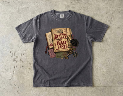 A dark gray crewneck t-shirt laid flat on a textured concrete surface, featuring a vintage-style graphic of a book titled 'GUILTY BAD TASTE' with a key, rose, and other old books.