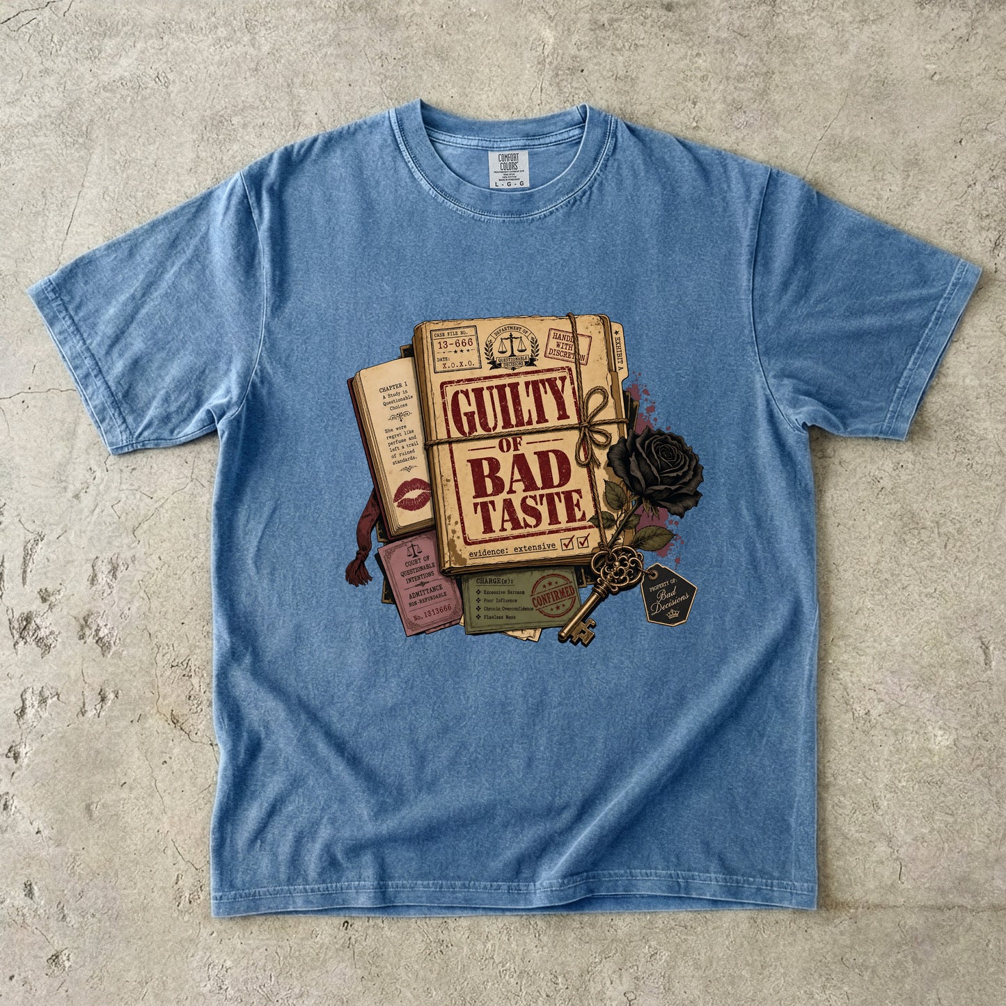 A light blue short-sleeve t-shirt laid flat on a textured concrete surface, featuring a vintage-style graphic print of an open book titled 'GUILTY OF BAD TASTE' with a key, rose, and lipstick marks.
