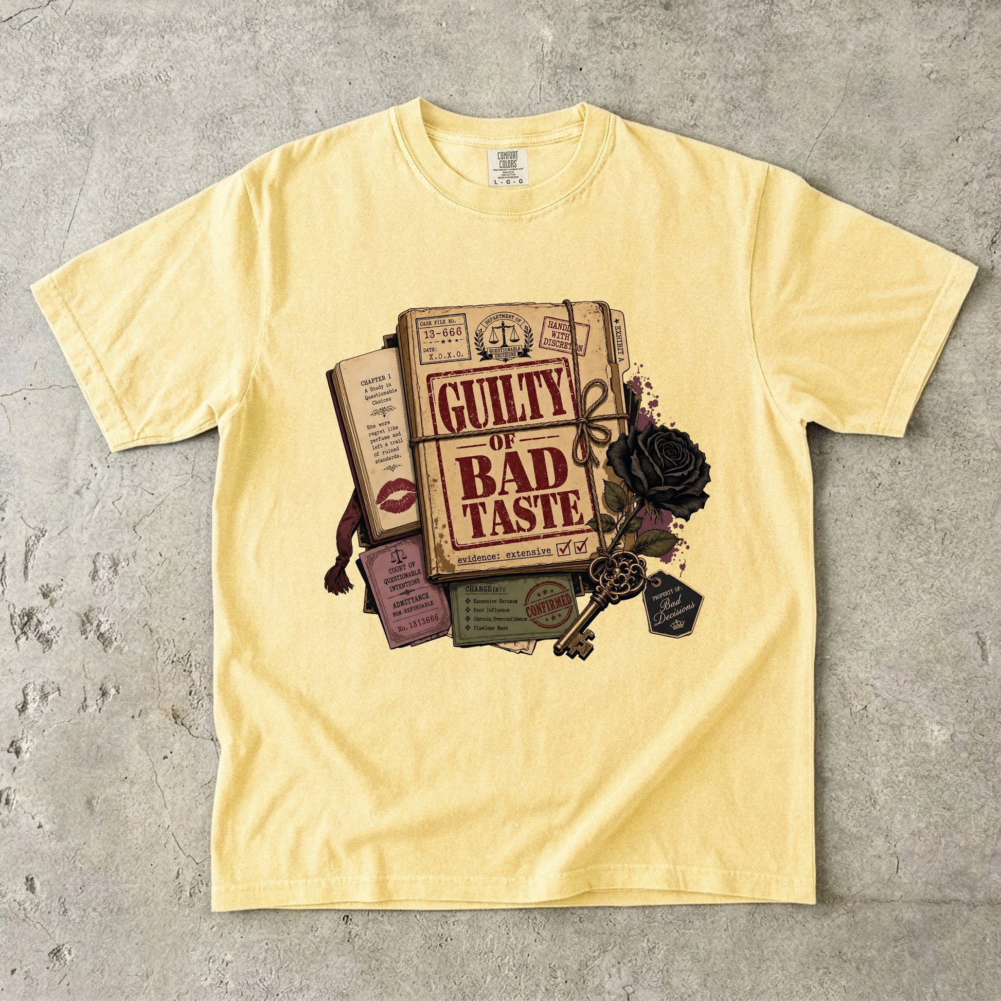 A pale yellow short-sleeve t-shirt laid flat on a textured gray concrete surface, featuring a vintage-style graphic print of a book titled 'GUILTY OF BAD TASTE' surrounded by a key, black rose, lipstick marks, and ticket details.