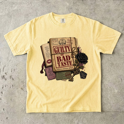 A pale yellow short-sleeve t-shirt laid flat on a textured gray concrete surface, featuring a vintage-style graphic print of a book titled 'GUILTY OF BAD TASTE' surrounded by a key, black rose, lipstick marks, and ticket details.