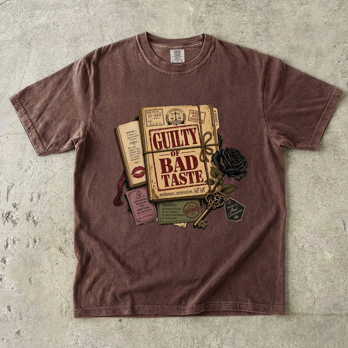A washed brown short-sleeve t-shirt laid flat on a textured gray concrete background, featuring a graphic print of a vintage book titled 'Guilty of Bad Taste' with a black rose, key, and ticket details.