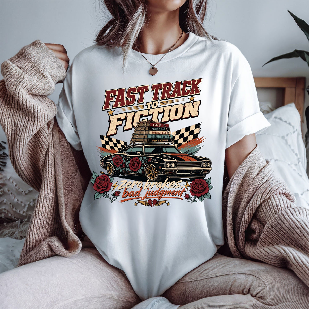 A woman wearing a white graphic t-shirt with a 'Fast Track to Fiction' car design, styled with a beige knit sweater draped off her shoulders in a cozy bedroom setting.