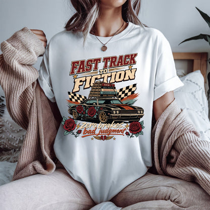 A woman wearing a white graphic t-shirt with a 'Fast Track to Fiction' car design, styled with a beige knit sweater draped off her shoulders in a cozy bedroom setting.