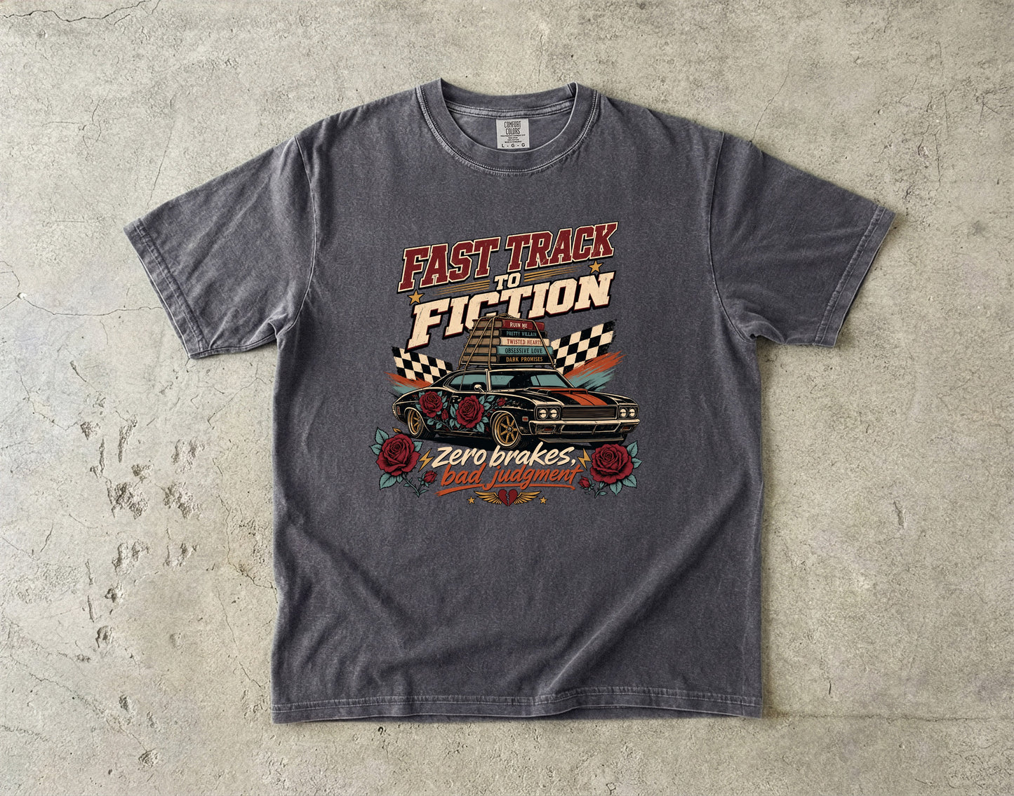 A dark gray graphic t-shirt laid flat on a textured concrete surface. The shirt features a retro-style print with the text 'FAST TRACK TO FICTION' and 'Zero brakes, gas filled!' surrounding an illustration of a classic race car, checkered flags, and roses.