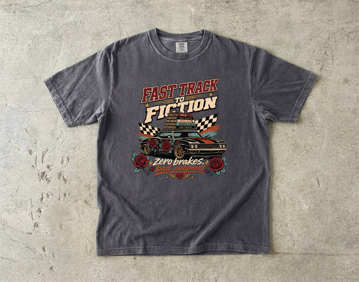 A dark gray graphic t-shirt laid flat on a textured concrete surface. The shirt features a retro-style print with the text 'FAST TRACK TO FICTION' and 'Zero brakes, gas filled!' surrounding an illustration of a classic race car, checkered flags, and roses.