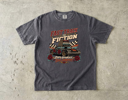 A dark gray graphic t-shirt laid flat on a textured concrete surface. The shirt features a retro-style print with the text 'FAST TRACK TO FICTION' and 'Zero brakes, gas filled!' surrounding an illustration of a classic race car, checkered flags, and roses.