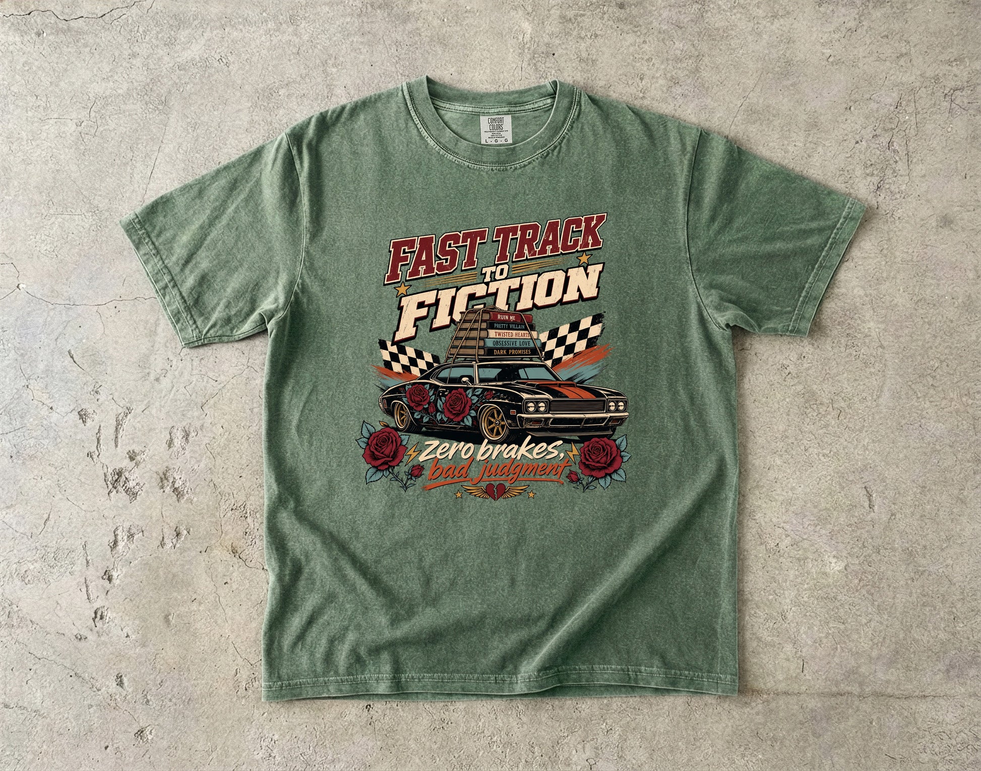 A green graphic t-shirt laid flat on a textured concrete surface. The shirt features a retro-style graphic with the text 'FAST TRACK TO FICTION' in red and cream, a black car with checkered flags, and 'Zero brakes bad attitudes' in red and cream.