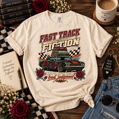 A cream-colored graphic t-shirt styled in a flat-lay arrangement on a dark wood background, surrounded by books, a mug, jeans, sunglasses, and flowers. The t-shirt features a retro graphic of a black car with a checkered flag and the text 'FAST TRACK TO FICTION' and 'ZERO TERRORISTES Dad Fugger'.