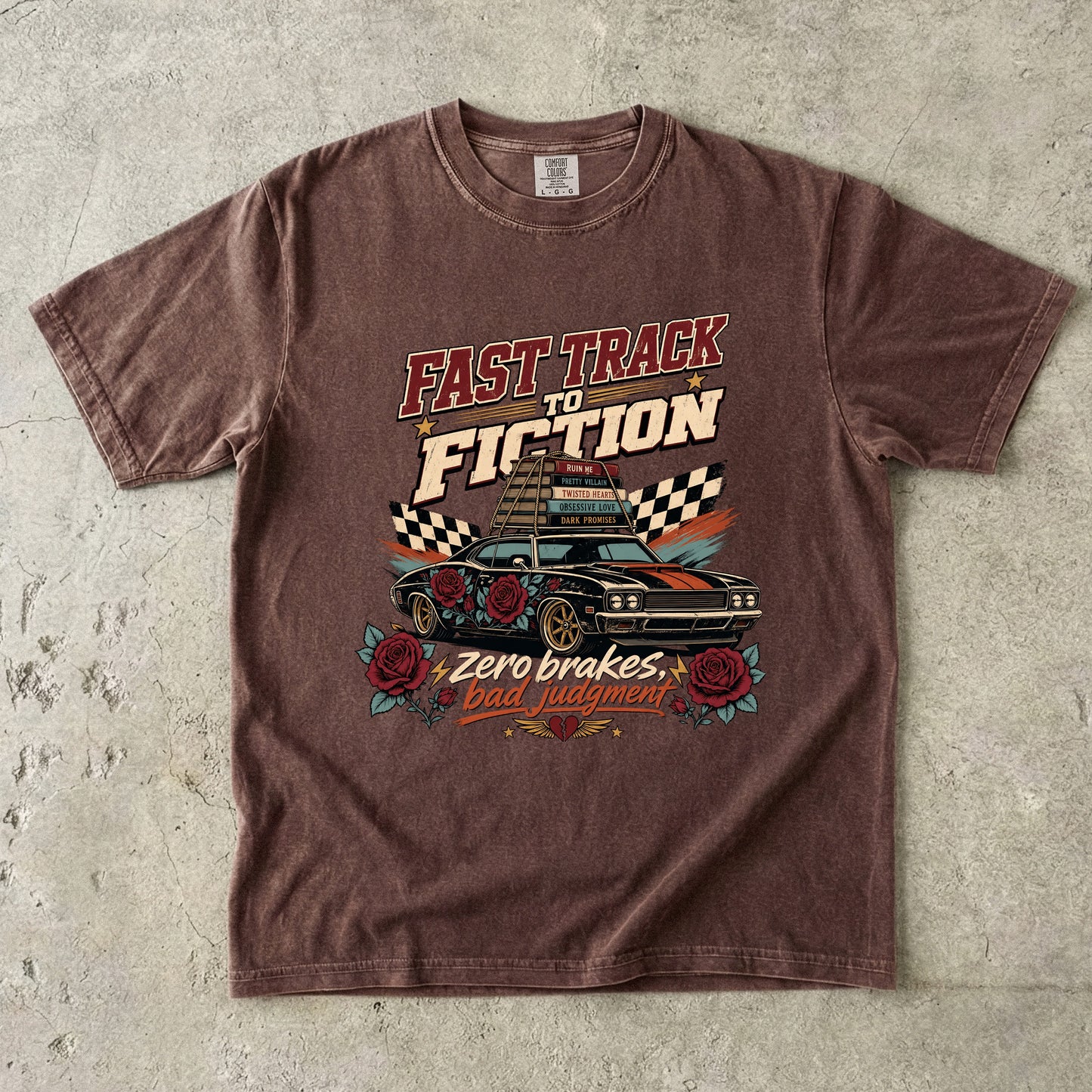 A brown short-sleeve graphic t-shirt laid flat on a textured gray concrete surface. The shirt features a distressed vintage-style graphic with the text 'FAST TRACK TO FICTION' and 'Zero brakes Bad judgment' surrounding an illustration of a race car, checkered flags, and roses.