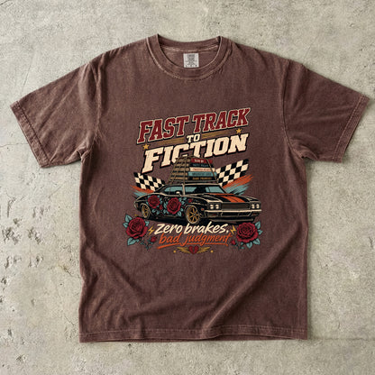A brown short-sleeve graphic t-shirt laid flat on a textured gray concrete surface. The shirt features a distressed vintage-style graphic with the text 'FAST TRACK TO FICTION' and 'Zero brakes Bad judgment' surrounding an illustration of a race car, checkered flags, and roses.