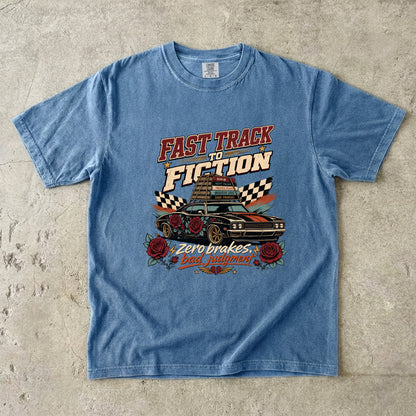 A light blue short-sleeve graphic t-shirt laid flat on a textured concrete surface. The graphic features a vintage-style car with roses and the text 'FAST TRACK TO FICTION' and 'Zero brakes.