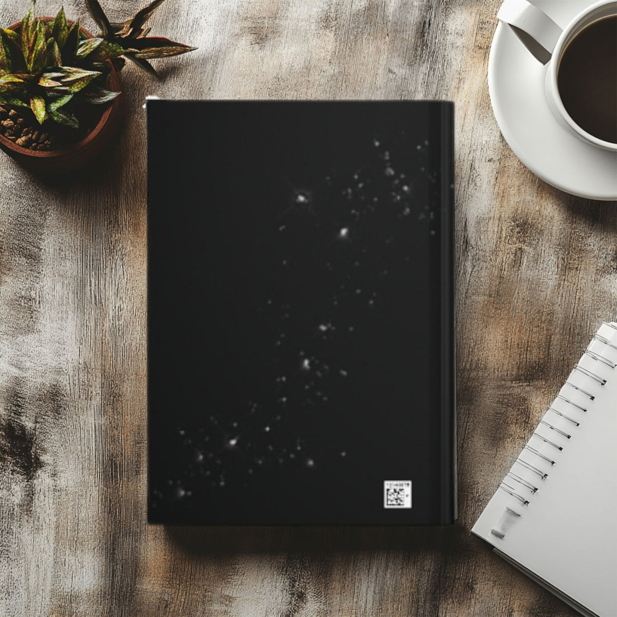 A black notebook with a white starry constellation pattern on the cover, styled in a flat-lay on a rustic wooden desk next to a coffee cup, a small potted succulent, and an open spiral notebook.