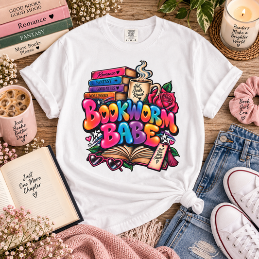 A white graphic T-shirt laid flat on a wooden surface, surrounded by styled props including books, a candle, jeans, sneakers, and a pink knit blanket. The shirt features a colorful illustrated design with stacked books, an open book, roses, hearts, and the text 'Bookworm Babes.