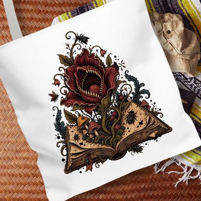 A white canvas tote bag featuring a detailed graphic print of a giant red flower with sharp teeth blooming from an open book, surrounded by mushrooms, spiders, and dark vines. The bag is styled on a brown woven mat with a striped blanket in the background.