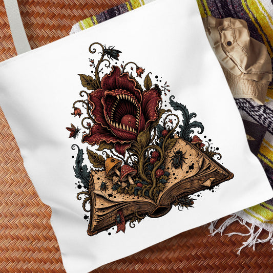 A white canvas tote bag featuring a detailed graphic print of a giant red flower with sharp teeth blooming from an open book, surrounded by mushrooms, spiders, and dark vines. The bag is styled on a brown woven mat with a striped blanket in the background.
