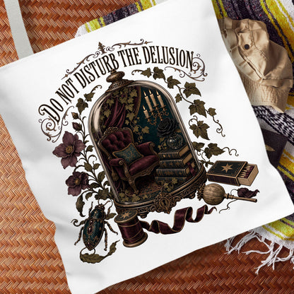 A white canvas tote bag featuring a detailed gothic illustration with the text 'DO NOT DISTURB THE DELUSION' and an ornate cloche containing a chair, books, and other mystical objects, displayed on a woven mat with a striped blanket in the background.