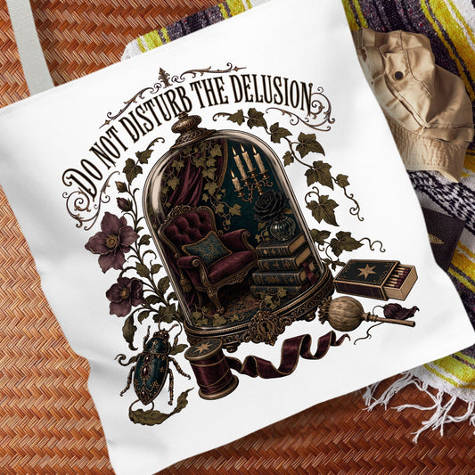 A white canvas tote bag featuring a detailed gothic illustration with the text 'DO NOT DISTURB THE DELUSION' and an ornate cloche containing a chair, books, and other mystical objects, displayed on a woven mat with a striped blanket in the background.