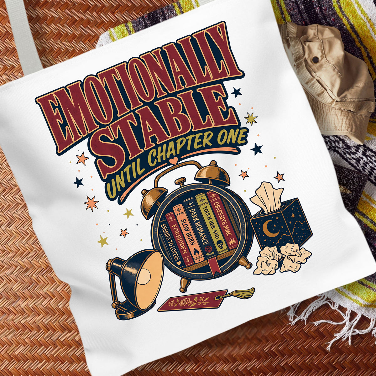 A white canvas tote bag with a graphic print that reads 'EMOTIONALLY STABLE UNTIL CHAPTER ONE' in large red and yellow lettering, surrounded by illustrations of a brass alarm clock, books, a lamp, tissue box, and crumpled paper. The bag is laid flat on a brown woven mat with a striped blanket in the background.