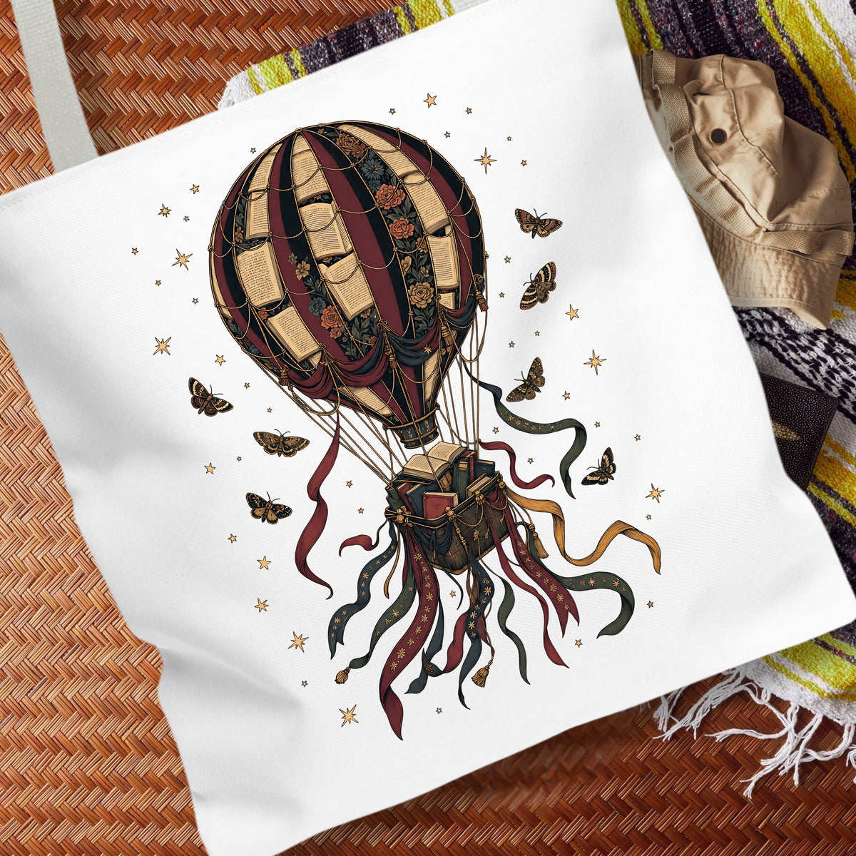 A white canvas tote bag featuring a detailed graphic of a vintage hot air balloon, butterflies, and stars, styled on a woven mat with a striped blanket in the background.
