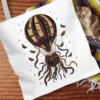 A white canvas tote bag featuring a detailed graphic of a vintage hot air balloon, butterflies, and stars, styled on a woven mat with a striped blanket in the background.