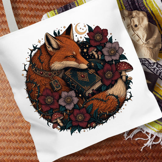 A white canvas tote bag featuring a detailed graphic illustration of a fox curled up with an open book, surrounded by flowers, mushrooms, and a crescent moon. The bag is displayed on a woven brown surface with a patterned blanket in the background.