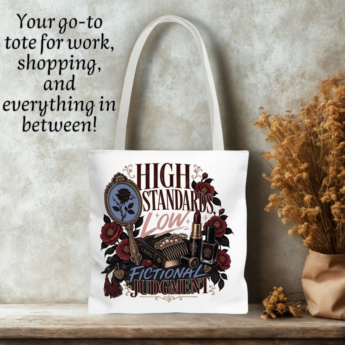 A white canvas tote bag with a vintage-style graphic reading 'HIGH STANDARDS LOW FICTIONAL JUDGMENT' in dark red and gold lettering, surrounded by illustrated roses, a mirror, and cosmetics. The bag is displayed on a wooden surface against a textured gray wall, with dried flowers and black decorative text on the left.