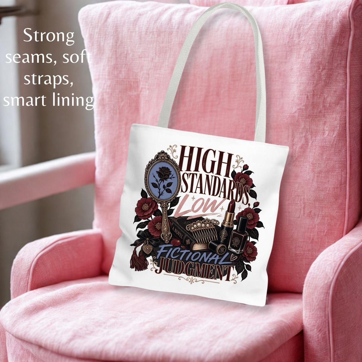 A white canvas tote bag with a graphic print of a mirror, lipstick, and roses, featuring the text 'HIGH STANDARDS Low FICTIONAL JUDGMENT'. The bag is displayed on a pink armchair with text overlays on the left describing its features.