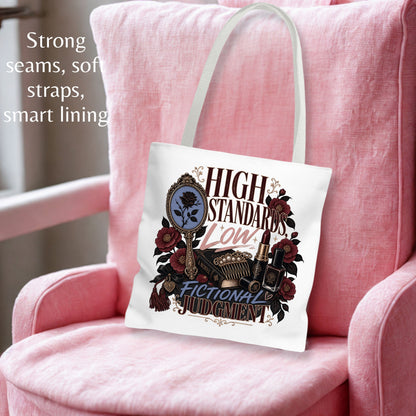 A white canvas tote bag with a graphic print of a mirror, lipstick, and roses, featuring the text 'HIGH STANDARDS Low FICTIONAL JUDGMENT'. The bag is displayed on a pink armchair with text overlays on the left describing its features.