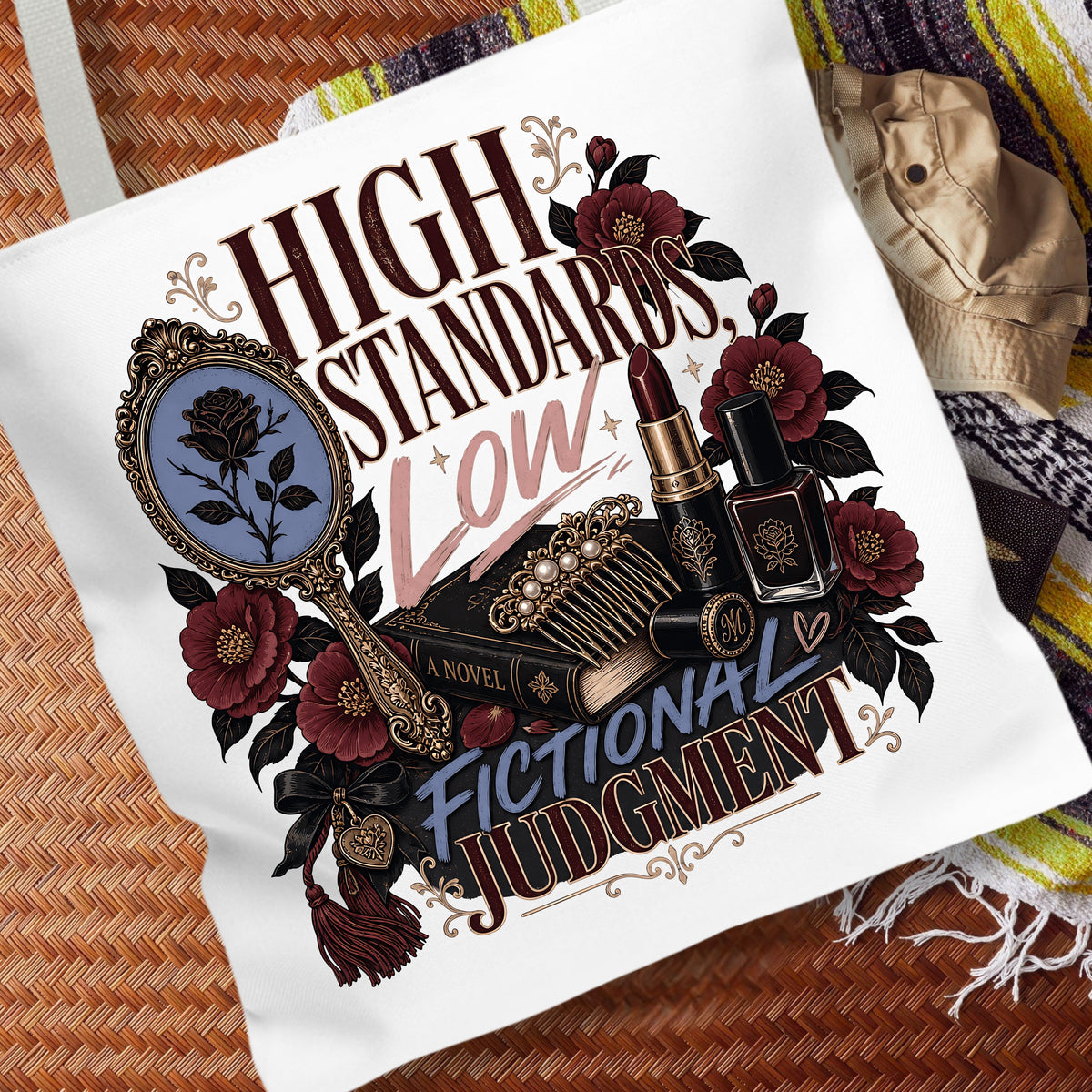 A white canvas tote bag featuring a gothic-themed graphic print with the text 'HIGH STANDARDS Low FICTIONAL JUDGMENT' and illustrated elements including a mirror, lipstick, perfume bottle, and roses, displayed on a woven brown mat with a striped blanket in the background.