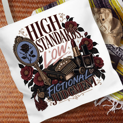 A white canvas tote bag featuring a gothic-themed graphic print with the text 'HIGH STANDARDS Low FICTIONAL JUDGMENT' and illustrated elements including a mirror, lipstick, perfume bottle, and roses, displayed on a woven brown mat with a striped blanket in the background.
