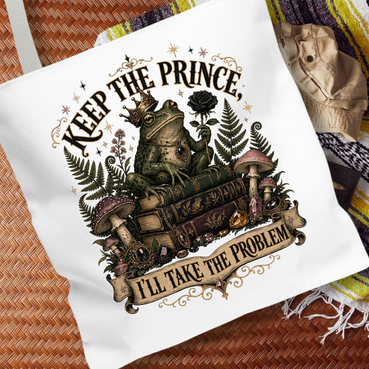 A white canvas tote bag featuring a whimsical graphic of a green frog wearing a gold crown, sitting on old books and holding a dark rose. The graphic includes the text 'KEEP THE PRINCE.