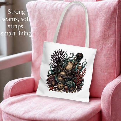 A white canvas tote bag with a detailed vintage-style ocean illustration, displayed on a pink armchair. The bag features a central graphic of a bottle, sea creatures, and the text 'LOST AT SEA.