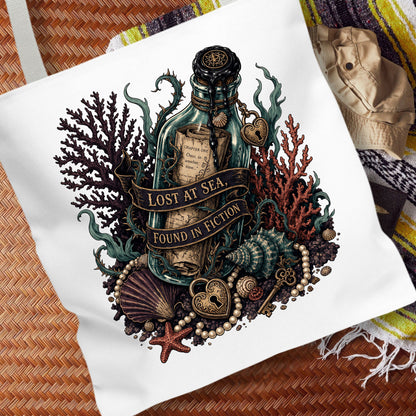 A white canvas tote bag featuring a detailed graphic print of a treasure bottle with the text 'LOST AT SEA. FOUND IN FICTION' surrounded by sea creatures, shells, pearls, and a key, styled on a woven mat with a striped blanket.