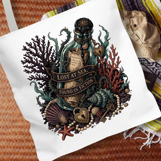 A white canvas tote bag featuring a detailed graphic print of a treasure bottle with the text 'LOST AT SEA. FOUND IN FICTION' surrounded by sea creatures, shells, pearls, and a key, styled on a woven mat with a striped blanket.